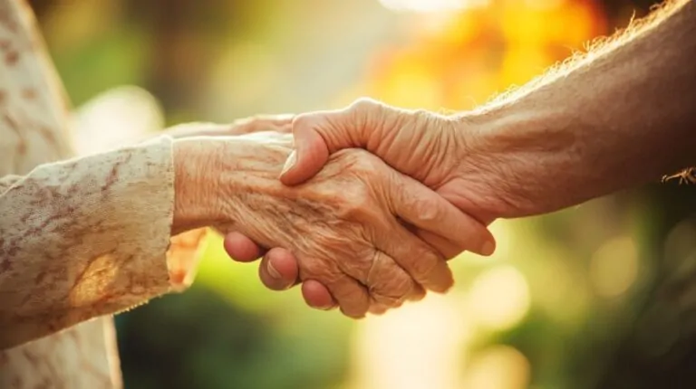 Two elderly hands gently holding each other in a warm outdoor setting, symbolizing support and understanding