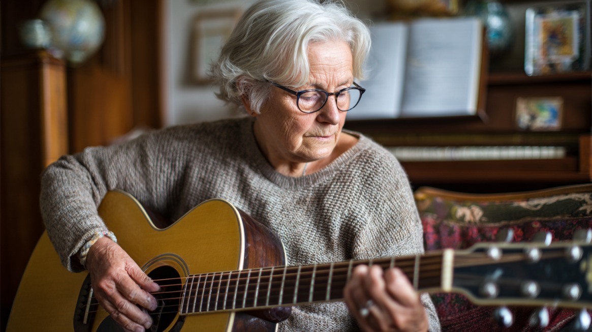 Older woman with white hair and glasses playing an acoustic guitar, sitting indoors in a cozy room with a piano in the background