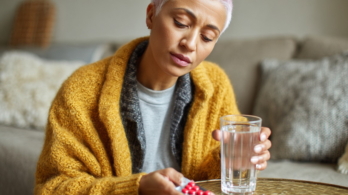 A woman holding a glass of water and examining a pack of pills while sitting on a couch