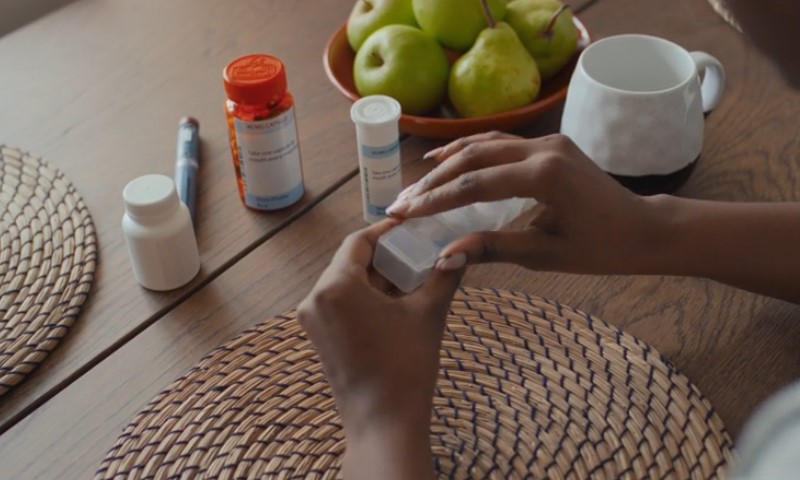 A person organizing pills in a weekly pill organizer on a wooden table with two woven placemats