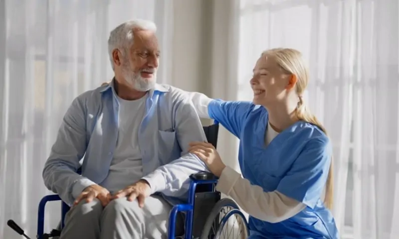 A smiling nurse in blue scrubs gently supports an elderly man sitting in a wheelchair