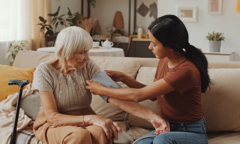 A young woman assists an elderly woman with a blood pressure cuff on a beige sofa