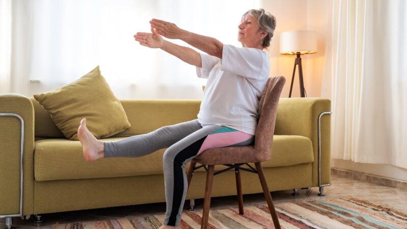 Older woman seated on a chair at home doing a gentle leg lift and arm stretch exercise