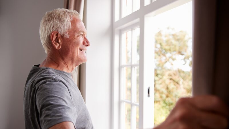Older man standing by a window looking outside at nature