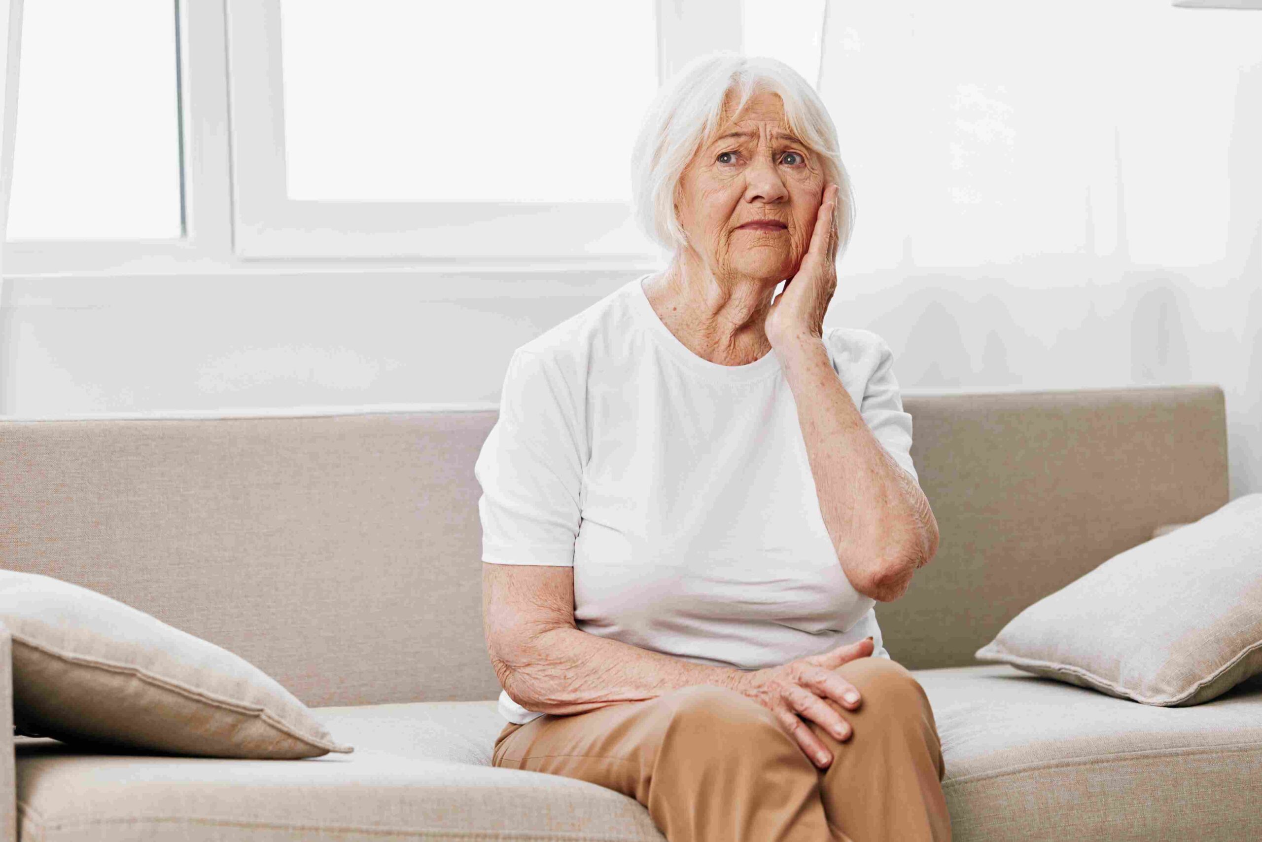 Elderly woman sitting on a couch holding her cheek in discomfort