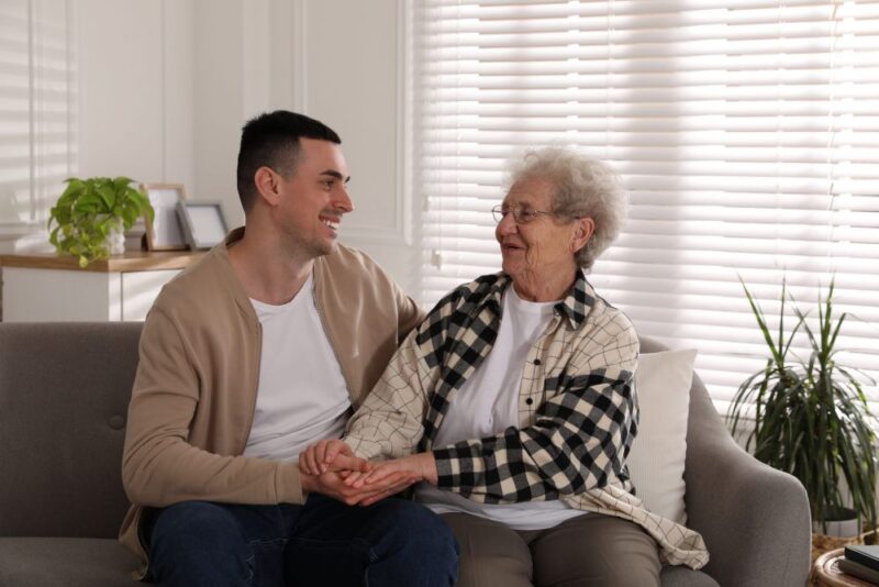 Young man sitting on a sofa with an elderly woman, smiling and holding her hands in a bright, comfortable living room